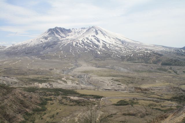 Panoramic view of Mount St. Helens with patches of snow on the peak and surrounding volcanic landscape. Great for use in travel and nature magazines, educational materials on geology and volcanoes, travel blogs, and promotional content for outdoor adventure tourism.
