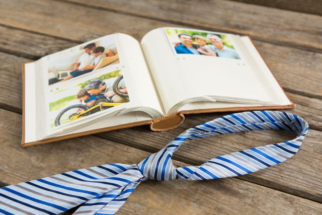 Open photo album filled with family photographs placed on rustic wooden table. A striped necktie lies across the table, adding a personal touch. Ideal for advertisements or articles on family traditions, celebrating memories, Father’s Day, or DIY photo books.