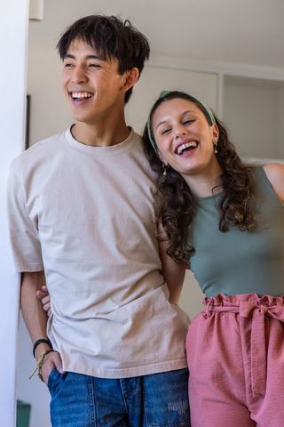 Laughing young couple leaning on doorway enjoying sunlit candid moment at home, casual