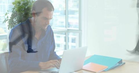 Focused man typing laptop at modern office desk overlooking cityscape with turquoise folder