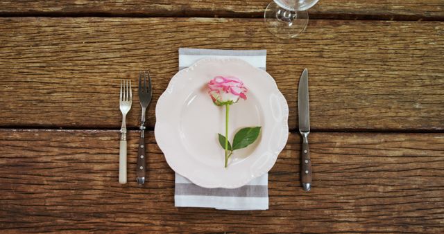 A pink flower adorns a white plate set with cutlery on a rustic wooden table, with copy space. The setting suggests a romantic or celebratory meal, with a focus on elegant simplicity.