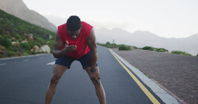Man wearing red sportswear is taking a break during his run on a mountain road. This image can be used to illustrate topics related to fitness, endurance, outdoor exercise, and the challenges of athletic training. It highlights the theme of persistence and the physical limits experienced during exercise.