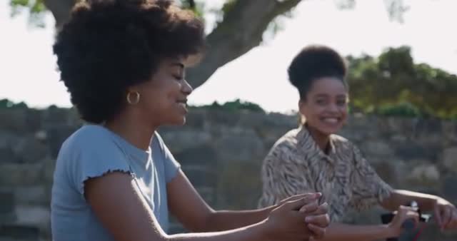 Two biracial women sitting and smiling at each other in an urban park with stone walls, trees, and clear skies, enjoying a sunny day. Ideal for themes related to leisure, friendship, outdoor activities, nature, wellbeing, positive connections, social interactions, and diverse communities.