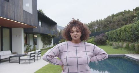 Curly-haired woman standing hands on hips by modern backyard pool wearing pink grid sweater