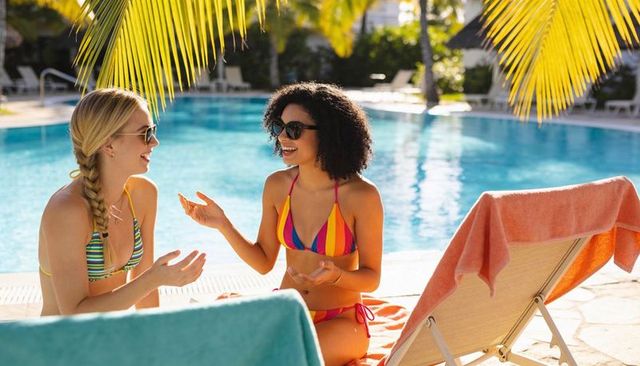 Relaxing friends chatting by tropical resort pool under palm fronds in colorful bikinis