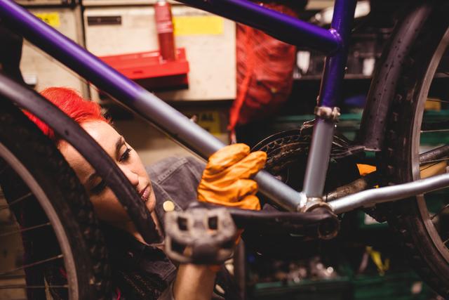 The image shows a female mechanic wearing orange gloves and repairing a bicycle in a workshop. She is focused on fixing the bike, highlighting her concentration and skill. The image can be used for promoting bike repair services, illustrating articles about women in traditionally male-dominated fields, or educational material about bicycle maintenance and repairs.