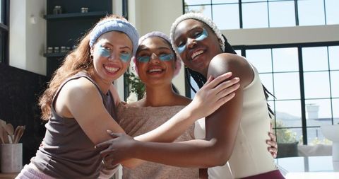 Hugging african american friends wearing teal under-eye patches smiling during home spa day