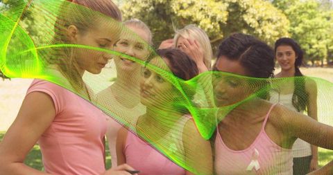 Group of women wearing pink ribbons in sunlit park holding smartphone with green digital overlay