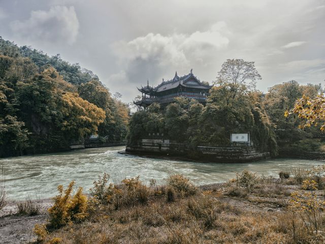 Ancient temple perched on a cliff beside a calm river enveloped by autumn foliage and cloudy sky. Can be used for topics related to history, architecture, Buddhist temples, tranquility, nature, and travel destinations, showcasing traditional structures and serene natural settings.