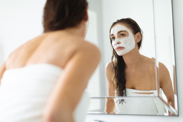 Woman with facial mask looking in bathroom mirror, wrapped in towel. Ideal for beauty, skincare, self-care, and wellness content. Useful for promoting spa services, skincare products, and relaxation routines.