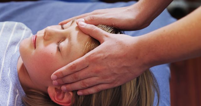 Child lying down on blue surface receiving head massage from therapist. Suitable for content related to child wellness, health care, therapy practices, spa and relaxation services, stress relief, and self-care routines. Useful in medical articles, wellness blogs, and promotional material for spa services.