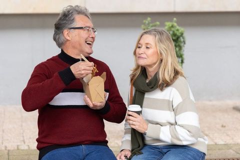 Senior couple enjoying takeout noodles and coffee on urban bench, relaxed outdoor living