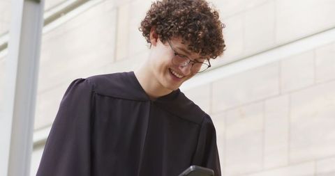 Young graduate checking phone in academic gown outside modern university facade