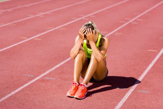 Female athlete sitting on running track, appearing upset and stressed. Ideal for use in articles or advertisements related to sports psychology, mental health in athletes, the pressures of competition, or fitness and training challenges.