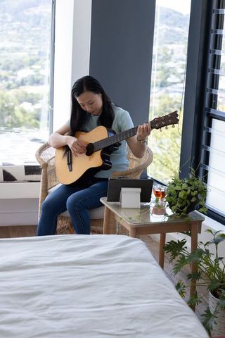 Asian woman practicing acoustic guitar in cozy sunlit bedroom with tablet for lessons
