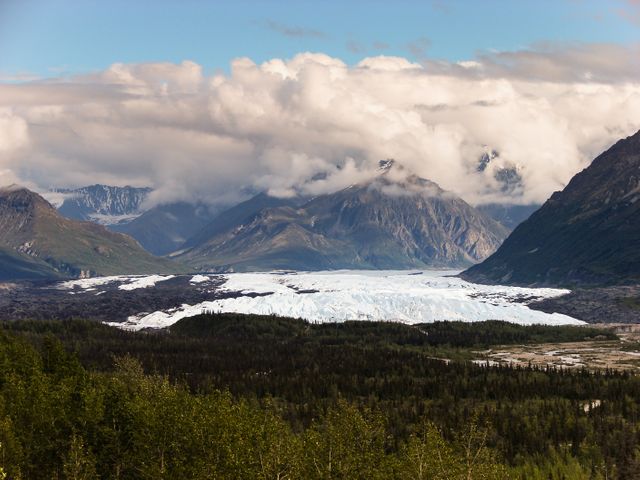 Picture showcasing a stunning glacier with surrounding mountain range under a cloudy sky. Snow-covered peaks rise in the distance, and dense forest stretches into the foreground. Ideal for travel brochures, adventure blogs, nature documentaries, and environmental awareness campaigns.