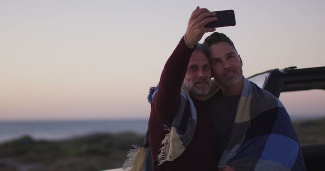 Happy caucasian gay male couple standing by car wearing blanket taking selfie at sunset on the beach. summer road trip and holiday in nature.