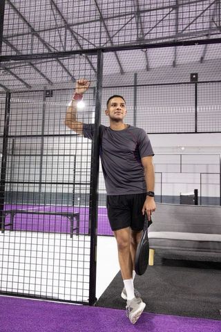 Young athlete leaning on mesh holding padel racket on purple indoor court