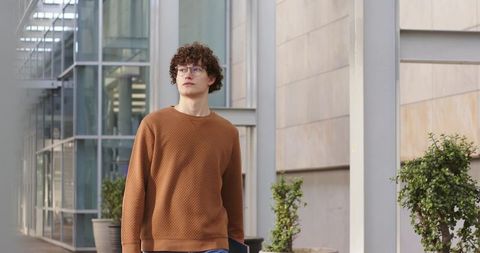 Curly-haired young man walking with laptop past modern glass building wearing brown sweater