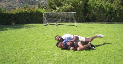 African american father and teenage son wrestling on sunny backyard lawn with portable soccer goal f