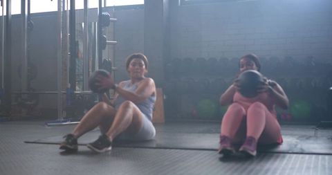 Diverse mid-adult women performing seated medicine ball twists in backlit gym for core strength