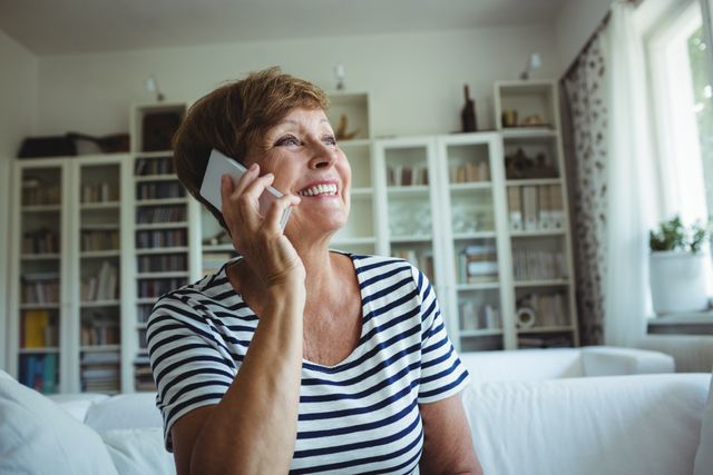 Senior woman smiling while talking on mobile phone in a cozy living room. Ideal for use in advertisements, articles, or websites related to senior lifestyle, communication technology, home living, and family connections.