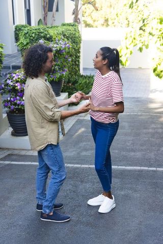 Couple holding hands and talking on sunlit driveway beside purple planters and greenery