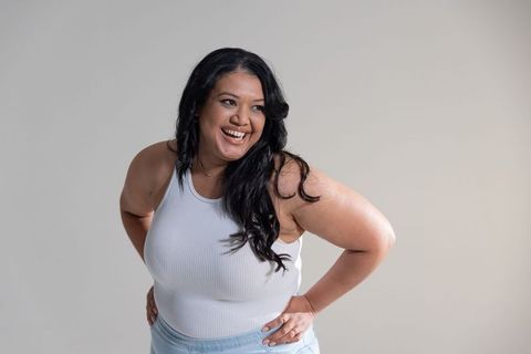 Confident plus-size woman smiling and leaning forward in white tank top and denim on neutral backdro