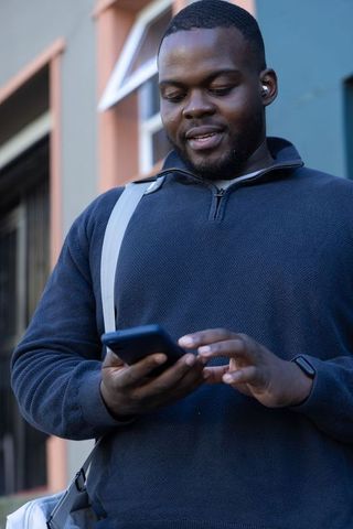 African american man using smartphone on urban sidewalk wearing sweater, earbud, smartwatch