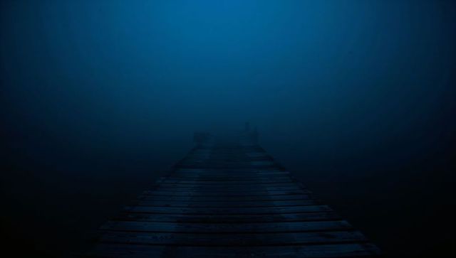 Weathered wooden pier leading into dense blue fog over lake at night, moody solitude