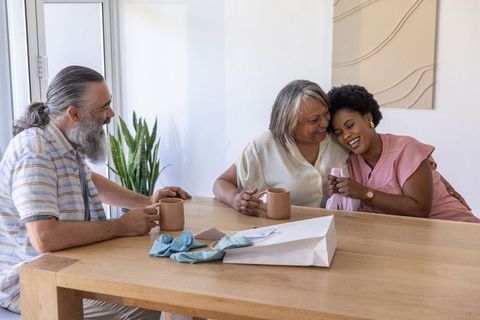 Multi-generational diverse family sharing hug while unwrapping gift over coffee at table