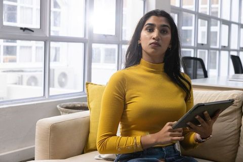 Indian woman sitting on beige sofa holding tablet in modern coworking lounge with windows