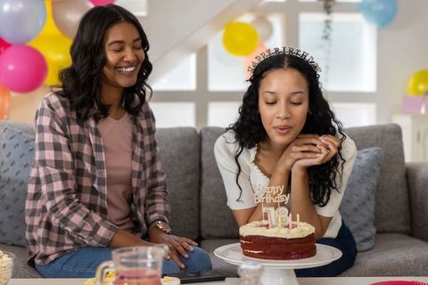 Diverse mother and teenage daughter celebrating 18th birthday with cake and tiara at home