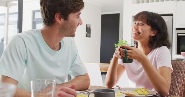 In this image, a couple is enjoying a relaxed breakfast together in a bright, modern kitchen. They seem to be involved in a pleasant conversation, creating a warm and friendly atmosphere. This stock photo can be ideal for promoting products and services related to relationships, kitchen appliances, home decor, or morning routines.