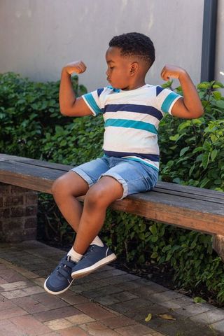Young african american boy flexing arms on bench in sunny courtyard