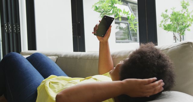 Image of plus size african american woman relaxing on sofa with smartphone. lifestyle, leisure, spending free time at home with technology.