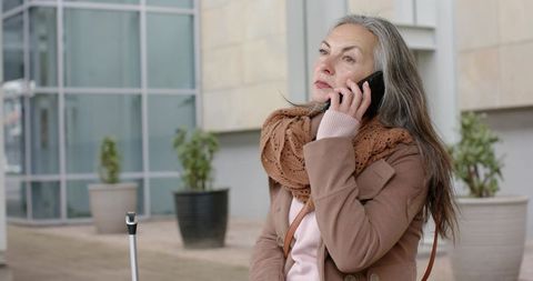 Senior woman traveling alone talking on phone waiting outside modern building with luggage