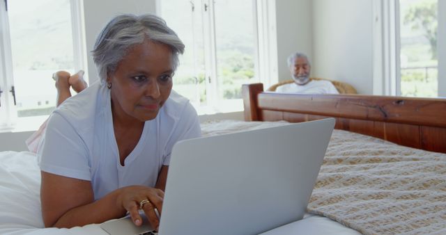 Elderly African American woman lying on bed, focused on using laptop. Light-filled room with another elderly person sitting in the background. Captures active senior lifestyle, modern technology use by older adults. Ideal for use in ads or articles about senior healthcare, retirement activities, technological literacy among seniors, comfortable living, or home environments for the elderly.