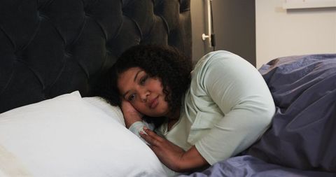 Woman lying on bed resting on pillow under gray duvet wearing mint top, calm cozy morning