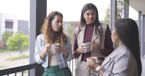 Diverse women colleagues sharing coffee and chatting on office balcony during break
