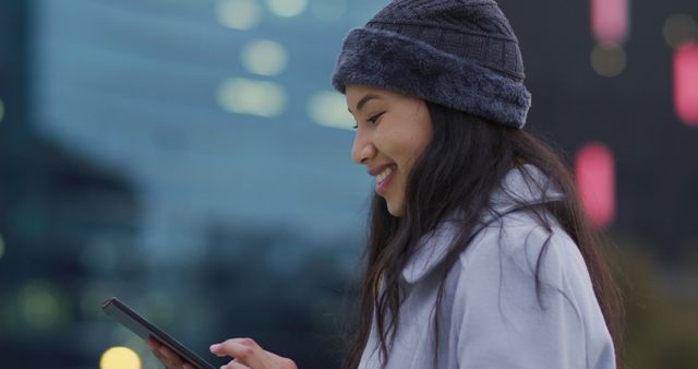 Young woman, dressed in a winter hat and coat, uses a smartphone while in an urban area with city lights in the background. Smiling and focused on her device, she demonstrates modern communication and lifestyle in a winter city. Ideal for illustrating themes of technology, connectivity, winter fashion, or urban living.