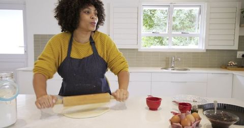 African american woman rolling dough on sunny kitchen island wearing apron and yellow sweater