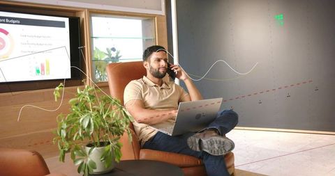 Remote worker talking on phone while working on laptop in modern office lounge