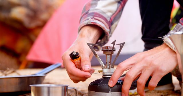 Focus is on hands lighting a portable camp stove with a lighter, with a tent and cooking utensils in the background. Useful for promoting outdoor gear, camping equipment, adventure travel, and nature experiences.