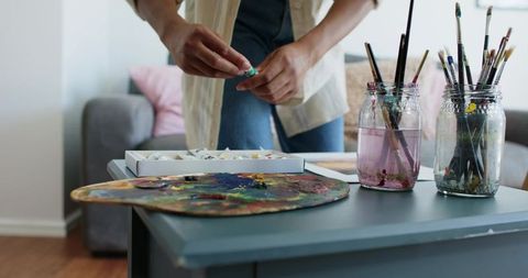 African american woman preparing paints on palette and brushes in cozy living room studio