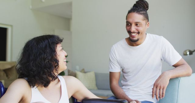 Happy biracial woman in wheelchair and male partner looking at each other and smiling in living room. wellbeing and domestic lifestyle with physical disability.