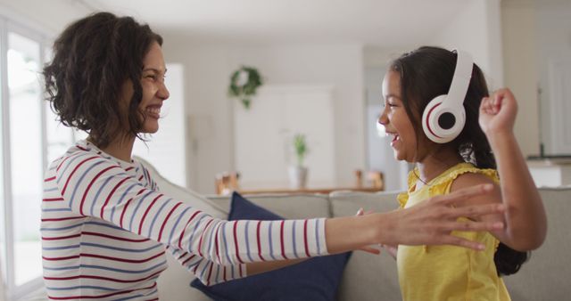 A mother and daughter sharing a joyful moment at home. They are sitting on a beige couch in a bright living room, wearing casual clothes. The daughter is excitedly wearing headphones, and both are extending arms towards each other, smiling. This image can be used for family-related content, advertising home audio products, or promoting parent-child bonding activities.