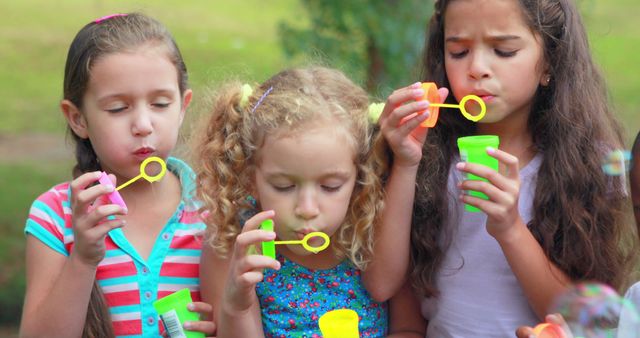 Three young girls are enjoying a playful moment blowing bubbles in an outdoor setting, with copy space. Their focused expressions and the floating bubbles add a sense of carefree joy to the image.