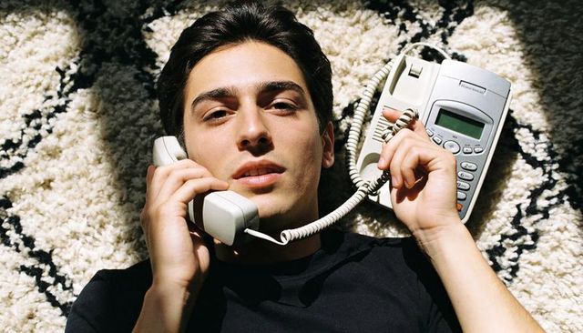 Young man lying on plush rug holding corded handset, talking on retro landline phone