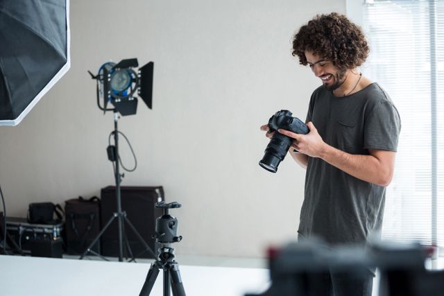 Male photographer with curly hair reviewing captured images on digital camera in a well-lit studio. Ideal for concepts related to photography, professional photographers, studio work, creative professions, and behind-the-scenes footage.